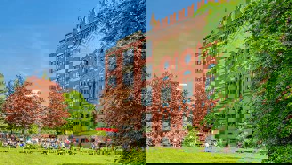 A large, ornate brick building next to a grassy area with people sitting at picnic tables under a sunny sky.