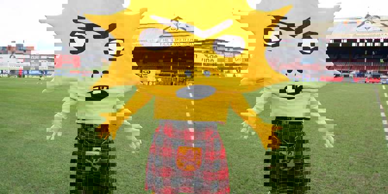 A person in a bright yellow, spiky sun-shaped mascot costume with a kilt and a small red hat stands on a grassy football pitch.