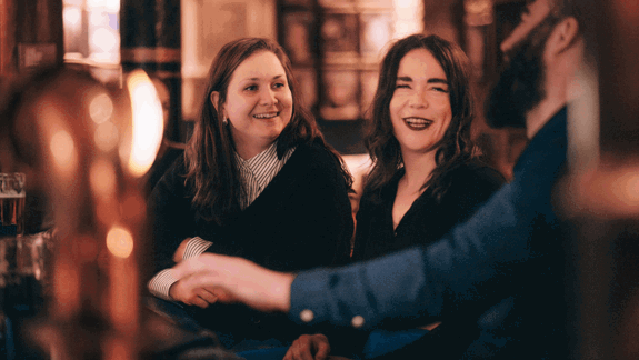 Group of 3 friends enjoying drinks at a cosy bar with warm lighting, traditional décor, and brass taps visible in foreground.