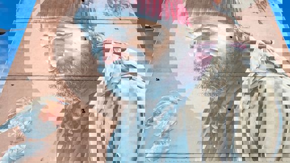 A low-angle shot captures a large mural of St Mungo in a red beanie and beige coat, with a robin perched on his finger. Another bird flies above his head.