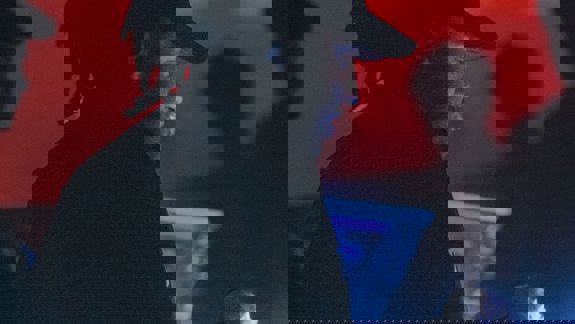 A DJ with dreadlocks and a baseball cap focuses on a mixing console in a dark club. Atmospheric red and blue lights glow in the background.