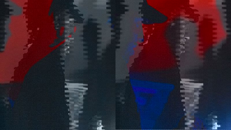 A DJ with dreadlocks and a baseball cap focuses on a mixing console in a dark club. Atmospheric red and blue lights glow in the background.