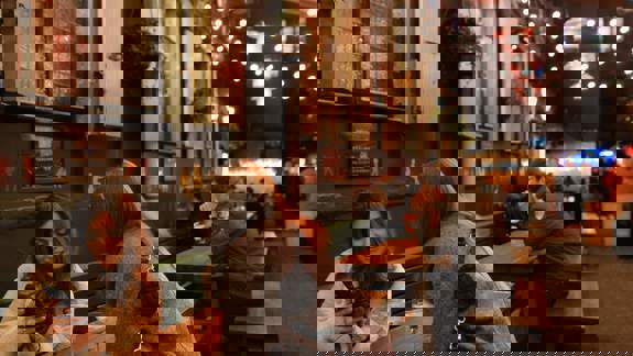 Cosy alley bar with wooden tables, string lights, hanging plants, and a festive crowd. Red pendant lights and shuttered windows add evening charm.