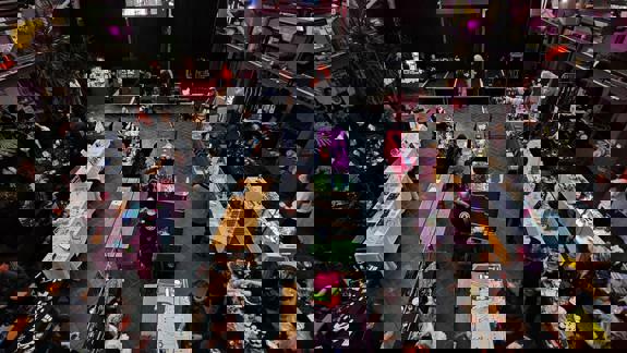 Indoor market with colourful stalls, benches, and shoppers, set in a modern venue with plants, stage lighting, and an elevated view of vendors selling art, accessories, and handmade goods.