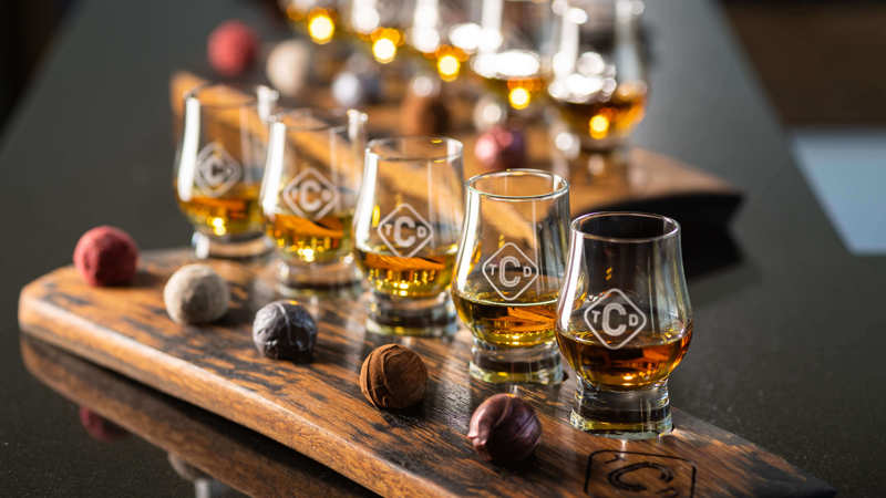 A row of six glasses of whisky on a wooden tray with small chocolate truffles scattered around them.