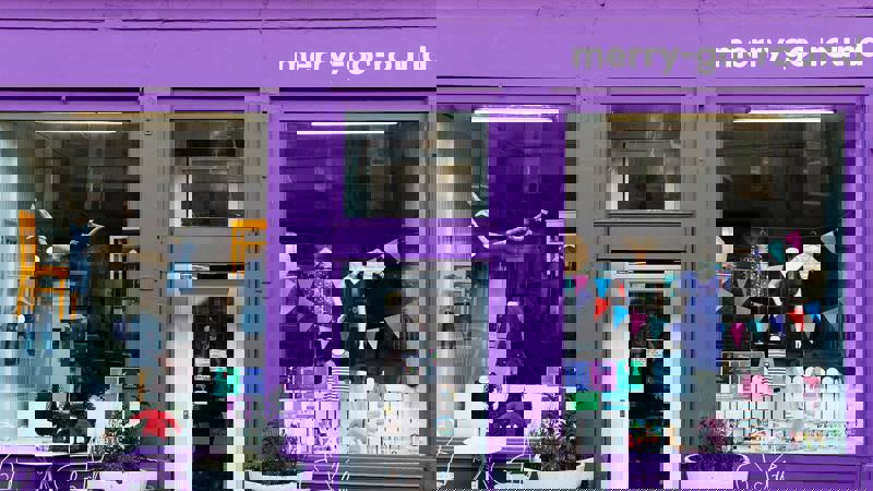 The storefront of merry-go-round, a children's clothing shop painted a vivid purple. Clothes are visible in the large window displays next to the main glass door.