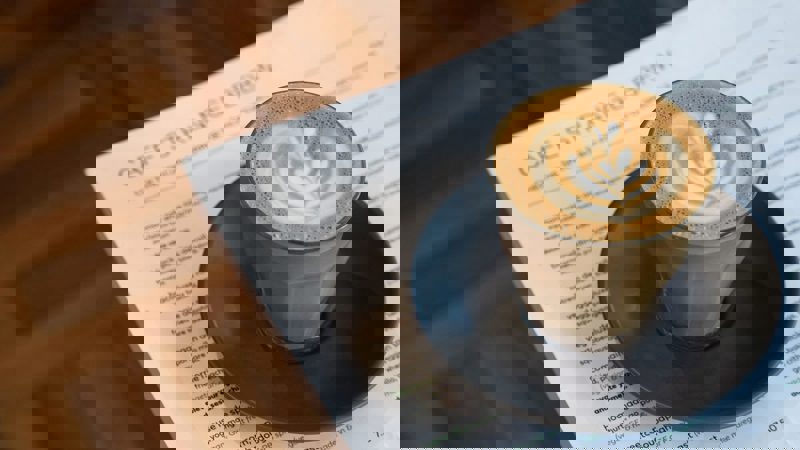 A flat lay shot of a coffee with latte art in a glass, placed on a black saucer. The coffee is sitting on top of a menu from Cafe Strange Brew, which lists various food items.