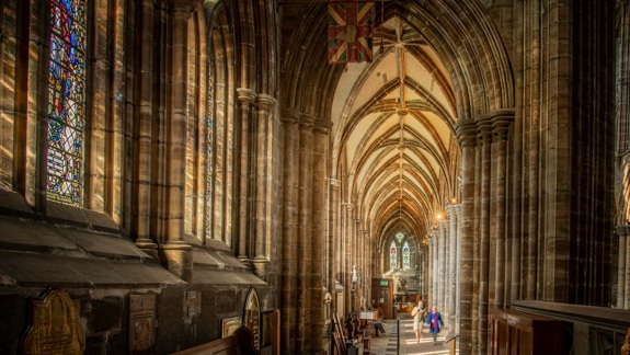 The interior of Glasgow Cathedral has grand arches where members of the public are walking through. Soft light filters through the stained glass windows.