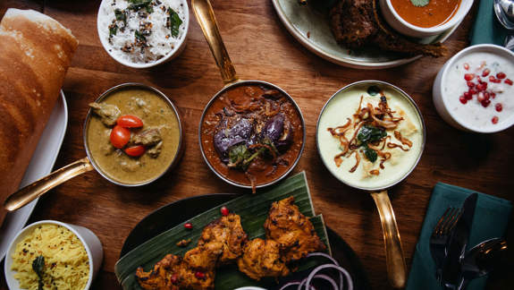 Assorted Southern Indian dishes including curries, rice, yogurt sides, grilled chicken on banana leaf, dosa, and fried onions, served with gold utensils and green napkins.