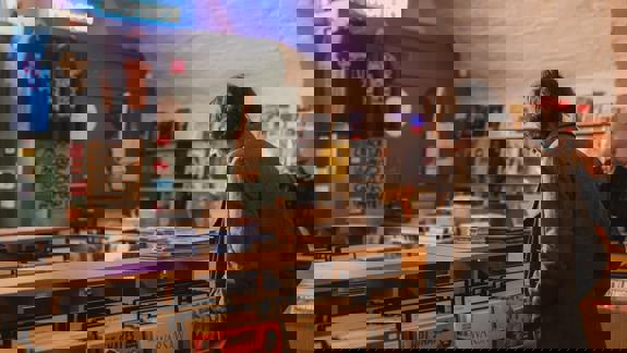 A person in a tweed jacket looks at a row of record albums in a store, with another person in the background.