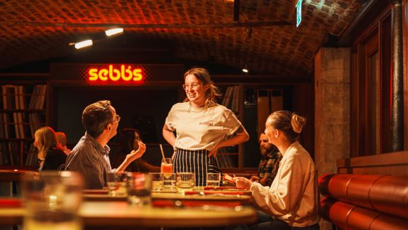 A smiling waitress talks to two seated customers in a warmly lit restaurant with a brick ceiling and a neon sign that reads “sebbs” in the background.