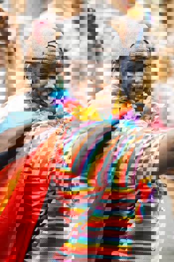 A smiling person wearing a rainbow-striped shirt, a Progress Pride flag like a cape, a lei, and a grey cap celebrates at a Pride event.