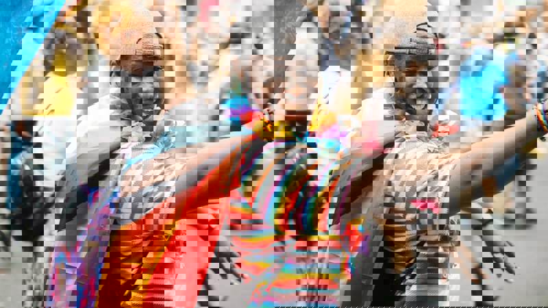 A smiling person wearing a rainbow-striped shirt, a Progress Pride flag like a cape, a lei, and a grey cap celebrates at a Pride event.