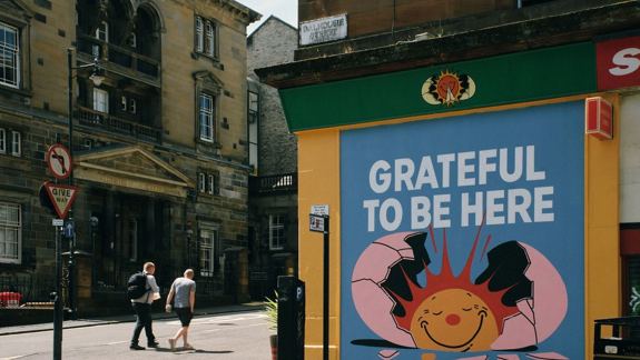 Street with two people walking past a colourful mural of a smiling sun breaking through a shell, and the words "Grateful to be here" in bold letters.