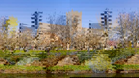 Paisley Abbey, a large historic stone church, stands beyond a river and a lush green park with blooming trees.