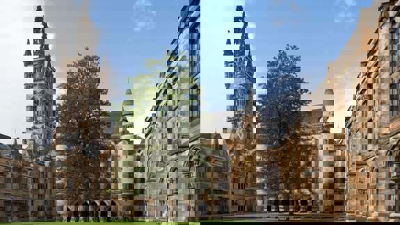 Historic stone buildings with a tall spire and a central courtyard with green grass.