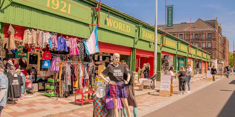 A street view of a market with mannequins and clothing racks outside a row of green and red shops.