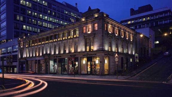 A well-lit corner building at dusk with light trails from passing cars in the foreground. The scene conveys a lively, urban atmosphere.