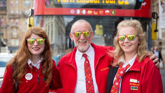 A smiling man in a red jacket and patterned tie stands between two young women, also in red jackets, all wearing reflective sunglasses, in front of a red tour bus.