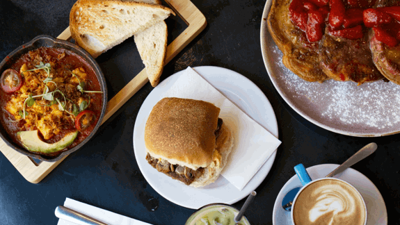Brunch spread with shakshuka in skillet, avocado, toast, sandwich, pancakes with strawberries, cappuccino with latte art, iced matcha latte, and silverware on dark table.