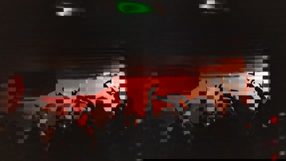 A crowd of people with their hands in the air, silhouetted against red and green lights in a dark music venue.