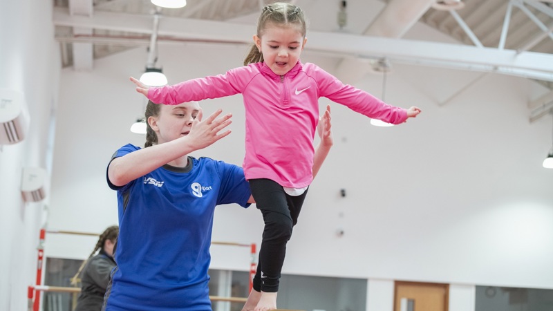 A young girl is balancing on a wooden balance beam with her arms out, with a female coach in a blue shirt standing beside her, ready to spot her.