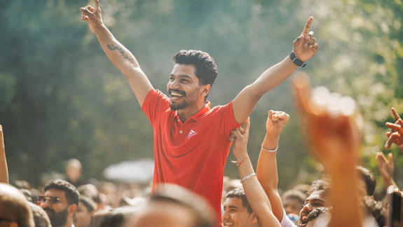 A man in a red polo shirt is held up by the crowd at an outdoor festival, smiling with his arms raised in a V-shape.