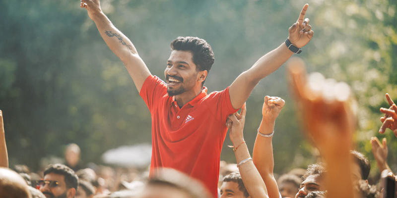 A man in a red polo shirt is held up by the crowd at an outdoor festival, smiling with his arms raised in a V-shape.