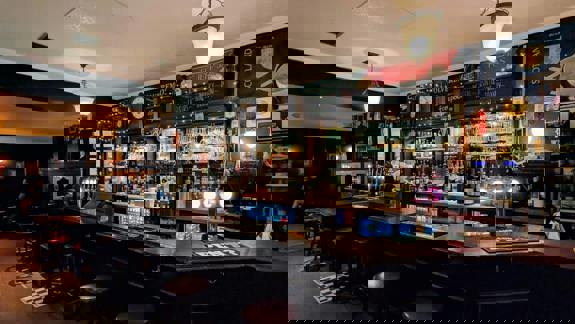 A warm, dimly lit traditional pub interior with a dark wood bar, bottles, glasses, and a Scottish flag display.