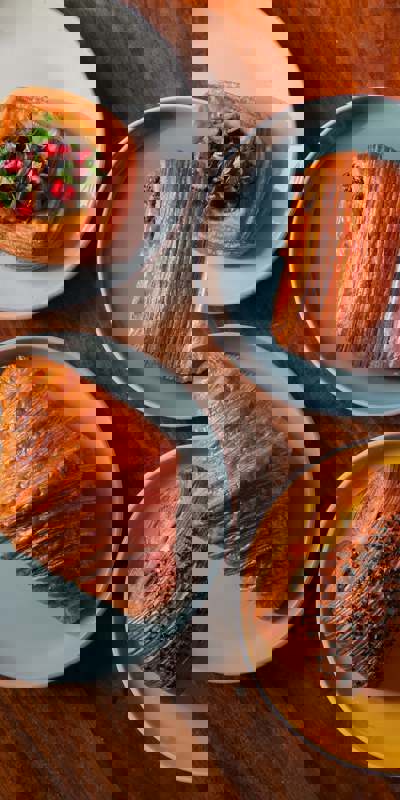 A wooden table holds four plates with pastries: a savoury danish with pomegranate, a chocolate pastry, a classic croissant, and a bun topped with seeds.