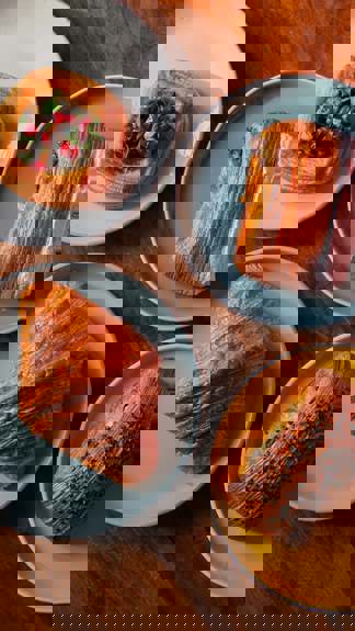 A wooden table holds four plates with pastries: a savoury danish with pomegranate, a chocolate pastry, a classic croissant, and a bun topped with seeds.