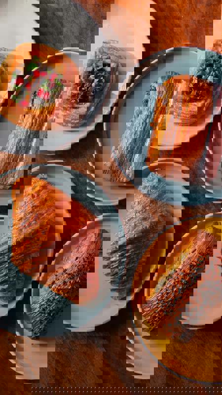 A wooden table holds four plates with pastries: a savoury danish with pomegranate, a chocolate pastry, a classic croissant, and a bun topped with seeds.