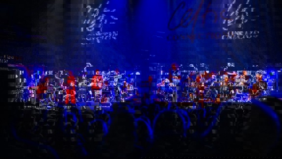 A wide shot of a band performing on a brightly lit stage under blue lights, with "Celtic Connections" projected in light above.