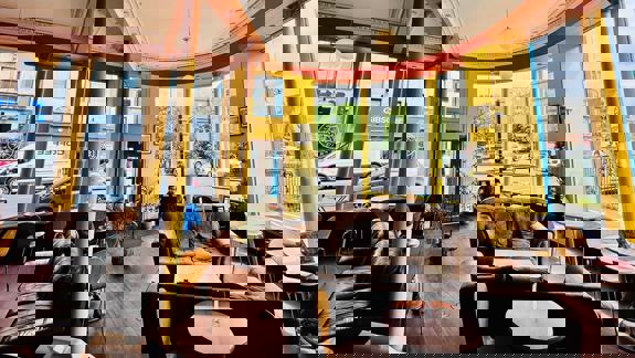 A cosy cafe interior with warm lighting, comfortable seating, and large windows looking out onto a street with cars and buildings.