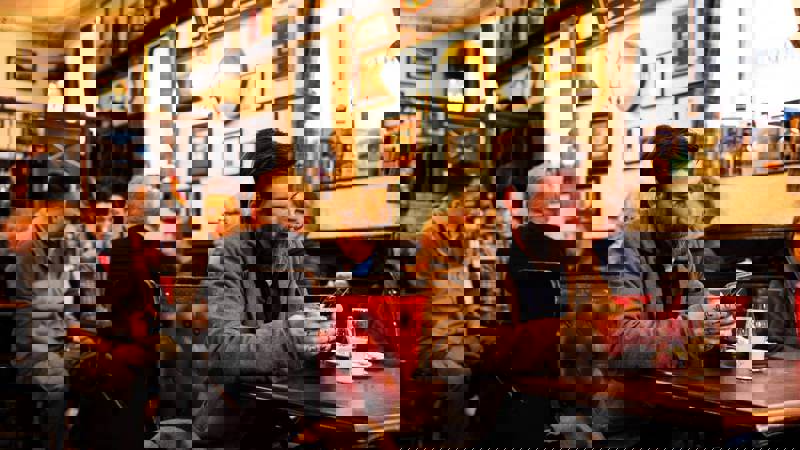 Customers relax inside The Pot Still, chatting or using phones in a warmly lit pub with red leather seating, framed decor, and whisky glasses on tables.