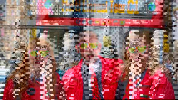 Three people, including an older person and two teenagers, stand smiling in front of a red city tour bus. They each wear matching sunglasses.