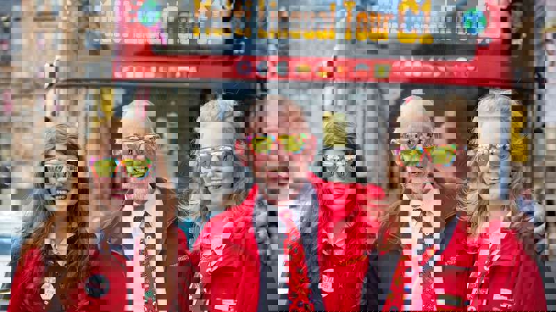 Three people, including an older person and two teenagers, stand smiling in front of a red city tour bus. They each wear matching sunglasses.