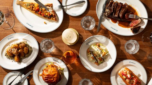 A wooden table set with several plated dishes, including a grilled fish, sliced steak, roasted potatoes, a burger, a salad, and a lobster roll, along with glasses, cutlery, and a lit candle