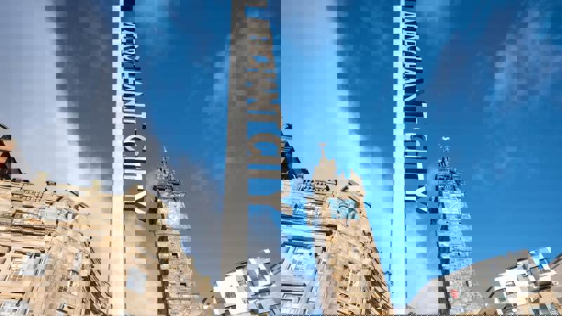 A tall, modern "MERCHANT CITY" sign against a blue sky, with historic stone buildings and a clock tower in the background, signifying the vibrant area.