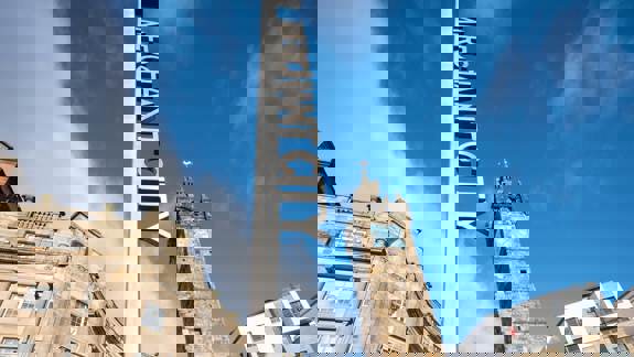 A tall, modern "MERCHANT CITY" sign against a blue sky, with historic stone buildings and a clock tower in the background, signifying the vibrant area.