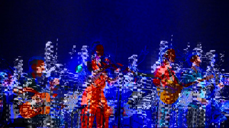 A music group performing on stage, featuring a woman playing a violin, a man playing a guitar, and another playing a bouzouki.