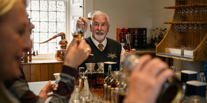 A distillery employee holds up a glass of whiskey. He is standing behind a bar with bottles and glasses. Two people are blurred in the foreground.