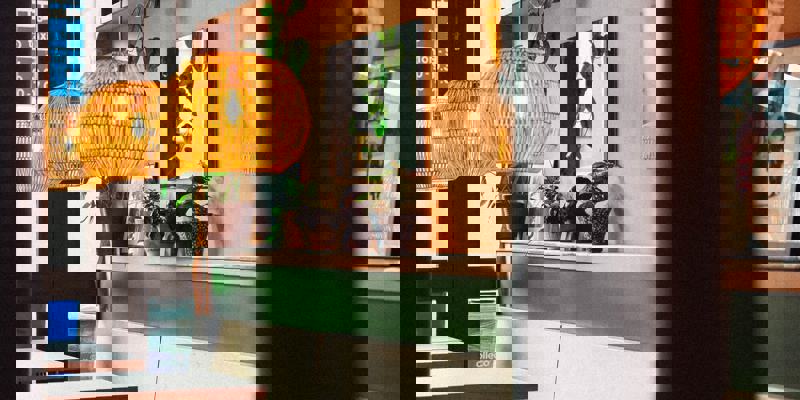 Wicker pendant lights hang above wooden tables in a cosy restaurant with plants on a shelf. A framed photo show a person walking.