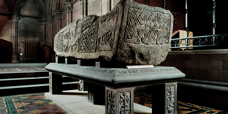 A large, carved stone sarcophagus with intricate knotwork patterns displayed on a plinth inside a historic church, lit by soft, natural light.
