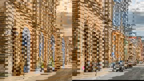 A sunlit street scene in Glasgow with a large, ornate building housing a Ralph Lauren store and traffic moving along the road.