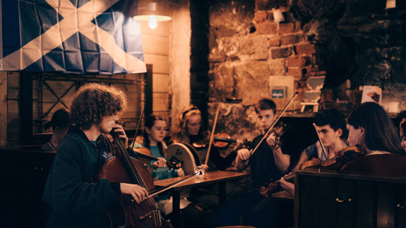 A group of young musicians plays traditional instruments in a cozy pub with a Scottish flag hanging on the wall.