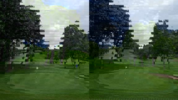 A view of the Cowglen Golf Club course in Glasgow, showing a neatly mown green with a flag, tree-lined fairways, and rolling hills under a partly cloudy sky.