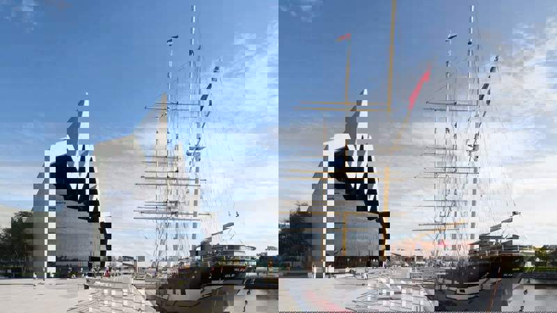 The modern, zig-zag shaped Riverside Museum next to the historic Tall Ship Glenlee docked on a river.