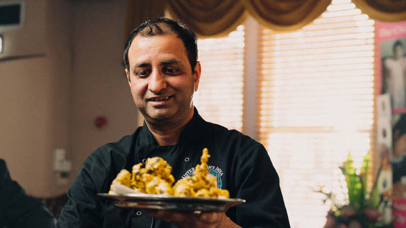 Chef in a black jacket smiling and holding a plate of Indian food.