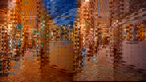 Twinkling fairy lights illuminate Ashton Lane, a charming cobblestone street lined with brick buildings, pubs,  and restaurants in Glasgow at dusk.